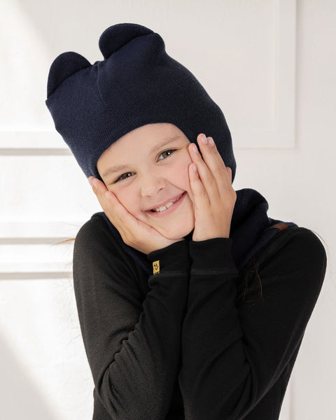 A girl holding her face with her palms, smiling, wearing the kids&#39; cotton balaclava with playful ears in dark blue color.