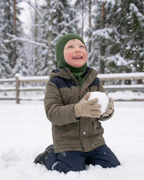 Smiling child dressed in winter outerwear and a green merino wool balaclava, playing with a snowball in a snowy forest setting with falling snowflakes.