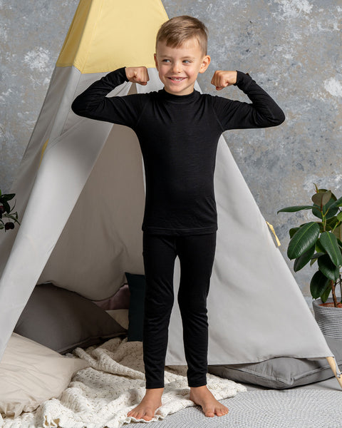 a boy wearing the Kids&#39; 160 Merino Pants in Black color. He has his arms raised up, acting strong. Behind him is a children&#39;s indoor play tent.