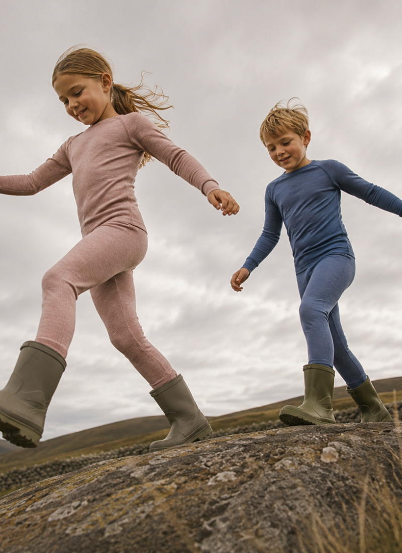 Two children outdoors on a rocky hillside under a cloudy sky, wearing matching long-sleeve leggings sets in dusty pink and blue, with olive green boots.