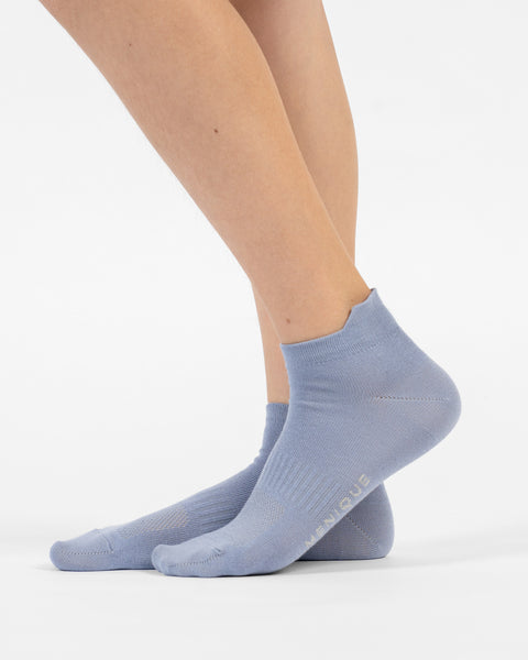 A side-profile view of a person's lower legs and feet wearing dusty blue cotton ankle socks, with the back foot slightly lifted to reveal branding on the sole.