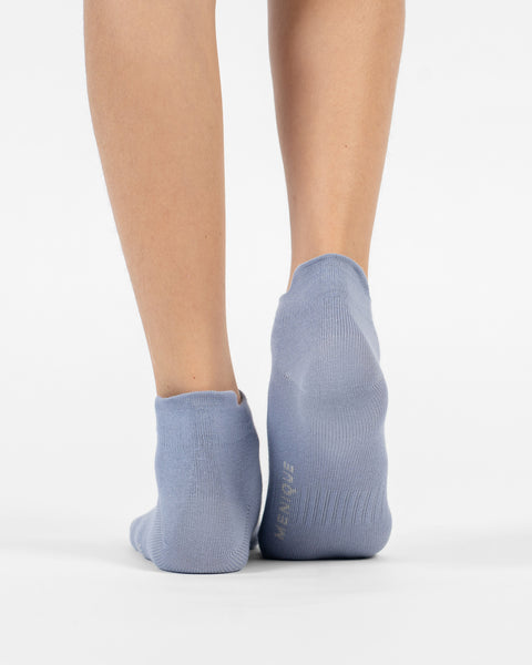 A back view of a person's lower legs and heels standing flat, wearing dusty blue cotton ankle socks with branding visible on the sole of the right foot.