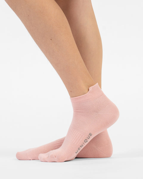 A side-profile view of a person's lower legs and feet wearing dusty pink cotton ankle socks, with the back foot slightly lifted to reveal branding on the sole.