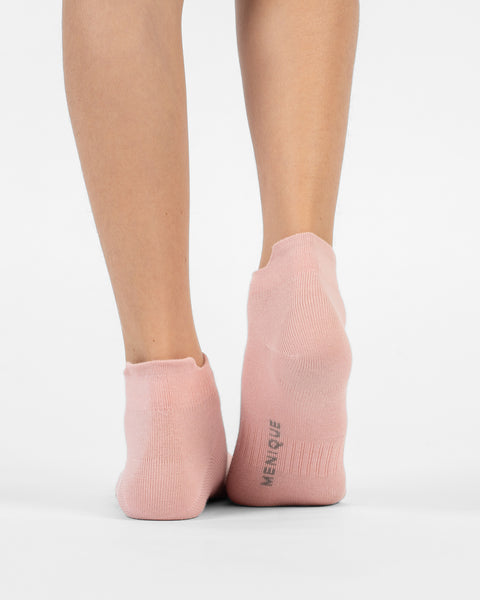A back view of a person's lower legs and heels standing flat, wearing dusty pink cotton ankle socks with branding visible on the sole of the right foot.