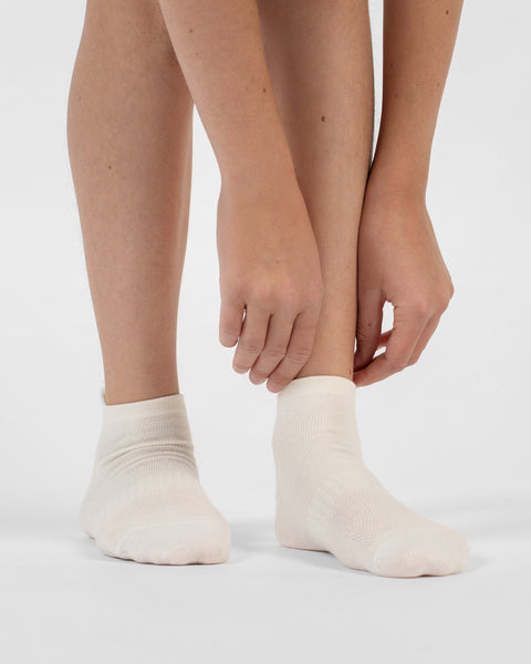 A front-facing view of a person's lower legs and feet, showing hands reaching down to adjust the cuff of a natural white cotton ankle sock.