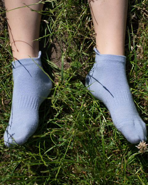 A top-down close-up view of a child's feet resting in a patch of green grass and dirt. The child is wearing light blue ankle socks.