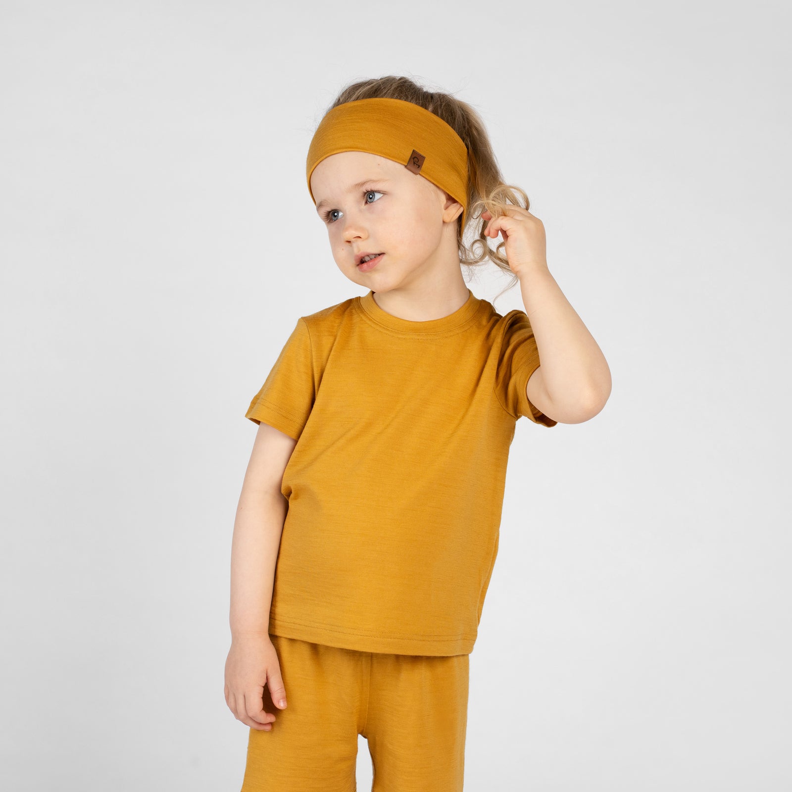 A studio portrait of a smiling young child looking off to the left against a plain light grey background. She is wearing a mustard yellow short-sleeved t-shirt and a matching wide mustard yellow headband with a small brown logo tag on the side, her hair pulled back in a ponytail.