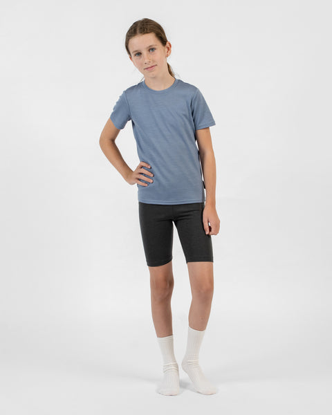 A young girl stands with her right hand on her hip, modeling charcoal merino cycling shorts and natural rib crew socks with a dusty blue short-sleeve top.