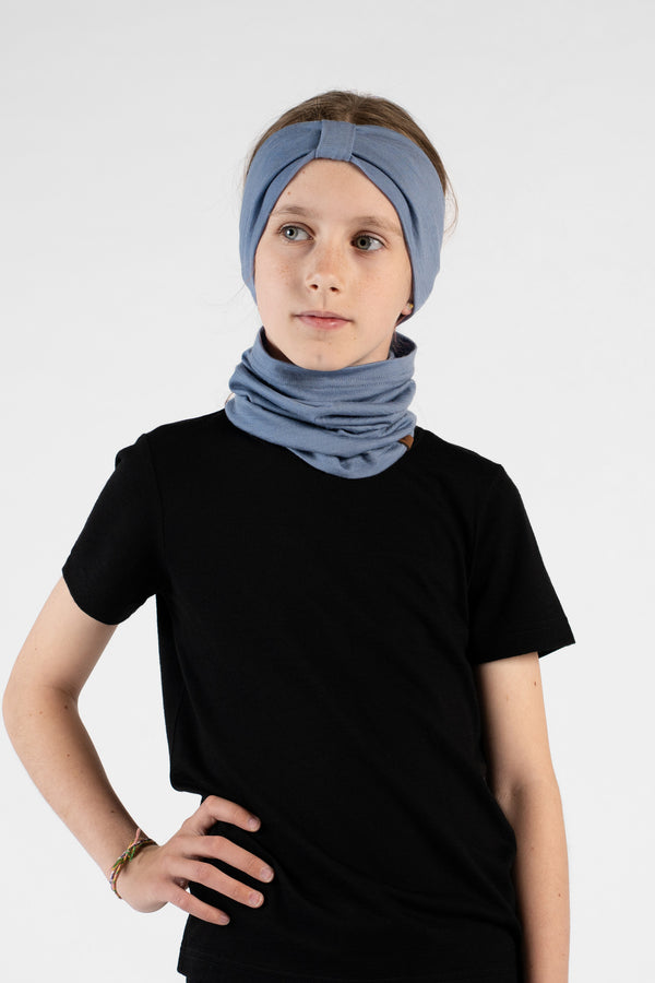 A studio portrait of a young girl standing with her hand on her hip, looking off to her left against a white background. She is wearing a black short-sleeved t-shirt, a dusty blue ribbon headband, and a matching dusty blue neck gaiter bunched loosely around her neck.
