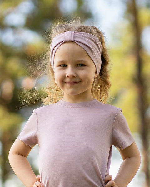 A young, fair-skinned girl with wavy blonde hair smiles gently at the camera while standing outdoors with her hands resting on her hips. She is wearing a light dusty pink short-sleeved t-shirt and a matching dusty pink fabric headband with a gathered, ribbon-like center detail. The background is a softly blurred outdoor setting with trees and dappled sunlight.