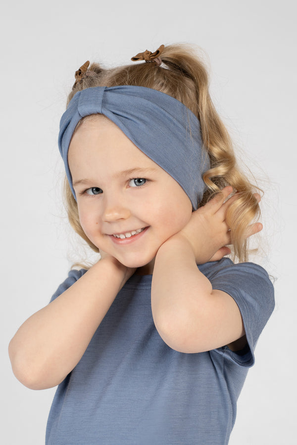 A close-up studio portrait of a young girl smiling directly at the camera. She wears a short-sleeved dusty blue top and a matching dusty blue ribbon headband. Her hands are gently resting behind her neck against a plain light grey background.
