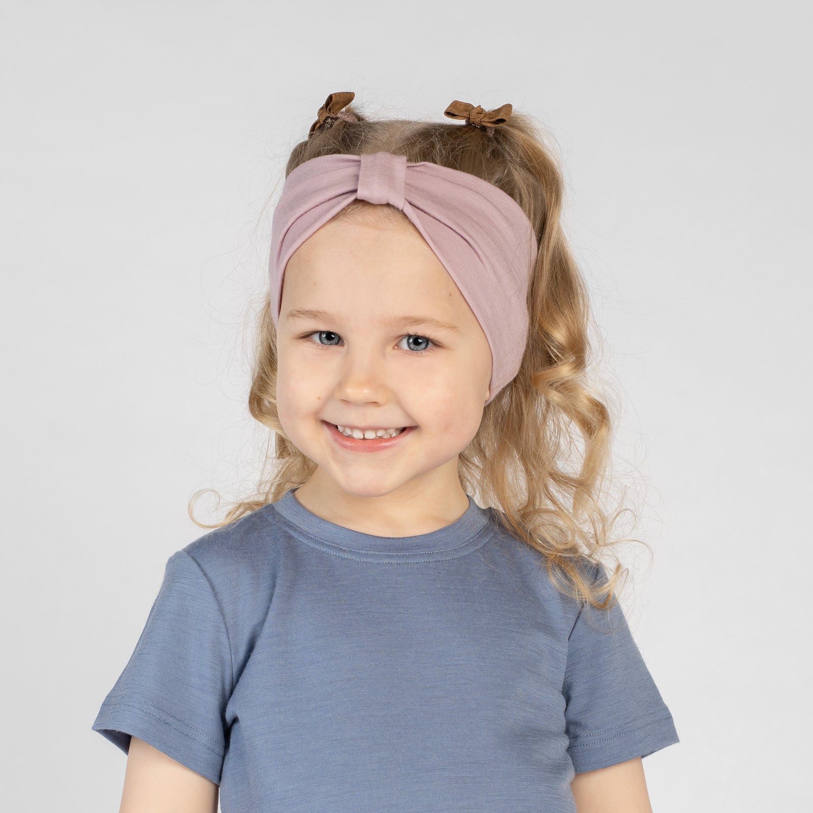 Young girl smiling outdoors wearing a dusty pink Merino wool T-shirt and a matching Merino wool headband. The soft, breathable accessories and clothing are showcased in a sunny natural setting, offering comfortable everyday wear for active ki
