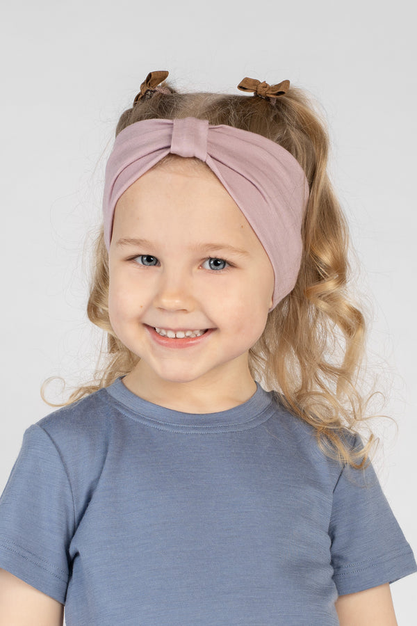 A close-up studio portrait of a young girl smiling directly at the camera. She is wearing a short-sleeved blue top and a dusty pink ribbon headband in her hair, against a plain light grey background.