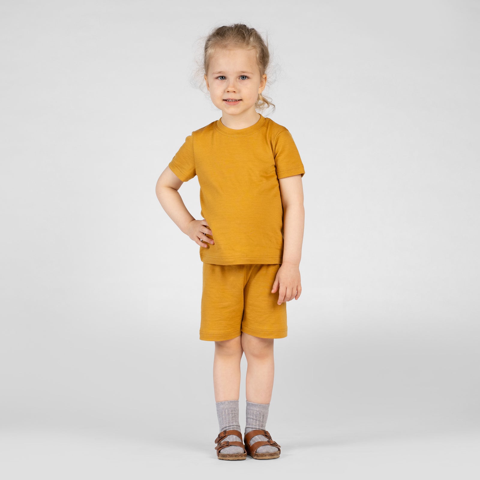 A close-up side profile view of a young child's torso and upper legs. The child is wearing a mustard yellow merino short-sleeve t-shirt and matching mustard yellow merino shorts. The child's hand is slightly lifting the hem of the t-shirt to show the elastic waistband and drawstring of the shorts.