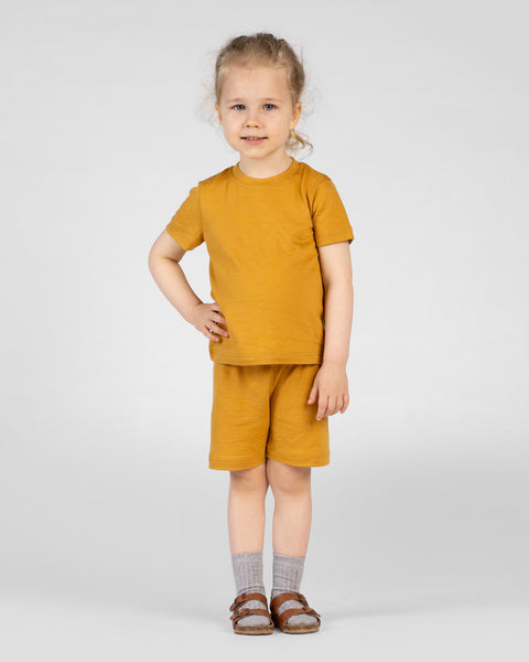 A full-body shot shows a child wearing a yellow short-sleeve t-shirt and matching shorts, paired with grey ribbed socks and brown buckled sandals. They are looking directly at the camera with a slight smile and one hand resting on their hip.
