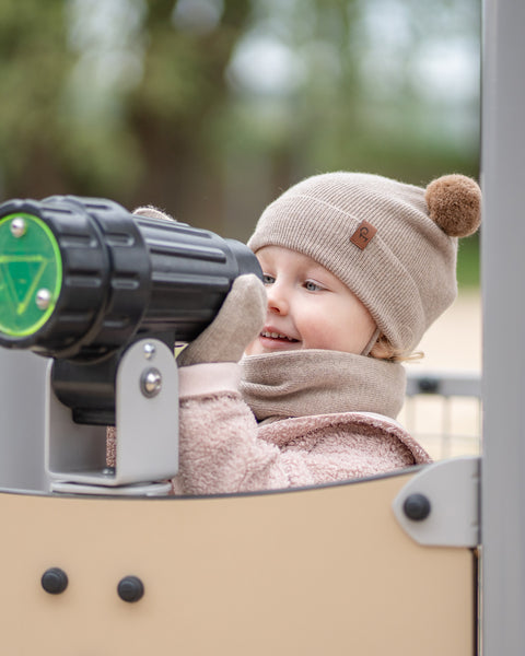A young child, wearing a creamy beige menique knit beanie with a brown pom-pom and a small brown tag, a matching menique neck warmer, gloves, and a pink textured jacket, is looking through a black toy telescope with green lenses on a playground structure. The child is smiling and looking to their right, seemingly engaged in play. The background is blurred with hints of green and brown.