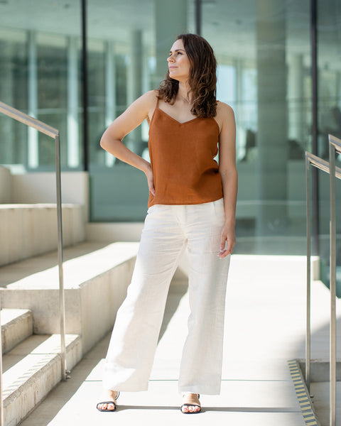A woman with brown hair wears a almond brown V-neck camisole top and white trousers, posing indoors in front of large glass windows.