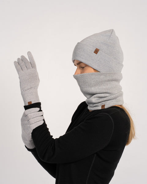 Woman wearing a light gray knit beanie, matching gloves, and a neck gaiter pulled up over the face, dressed in a black long-sleeve top, shown in profile with one hand raised against a plain background.