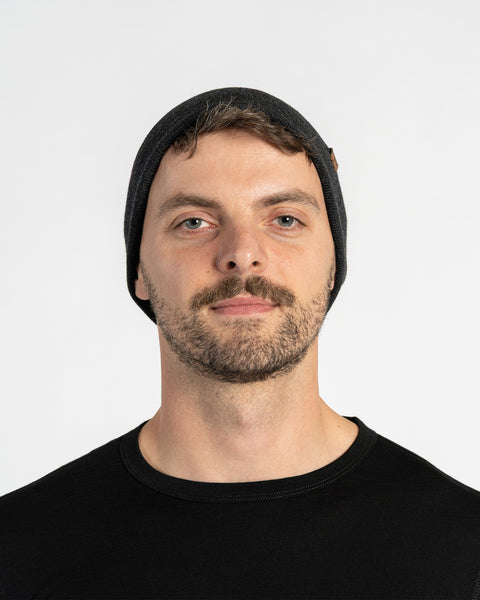 A man with light skin, blue eyes, and a beard is looking forward. He wears a dark gray colored menique knit beanie with a small brown patch on the side, and a black crew-neck shirt.