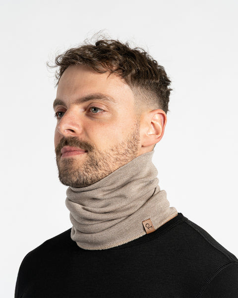 A man with short dark hair and a neatly trimmed beard models a creamy beige, knitted neck gaiter. He is wearing a black shirt and is pictured from the chest up, looking away from the camera against a white background.