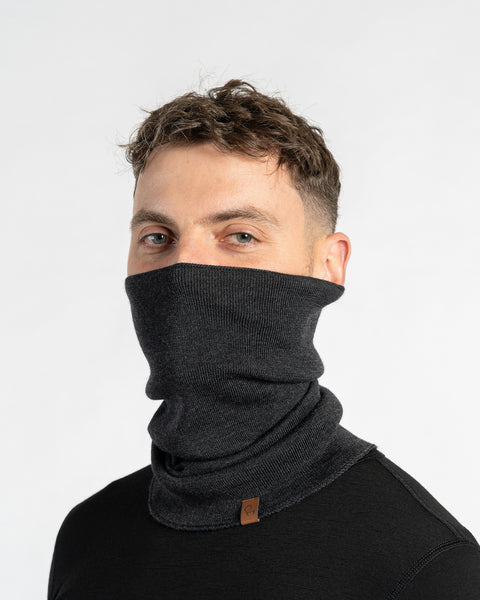 A man with short brown hair and light-colored eyes looks at the camera while modeling a dark gray knitted neck gaiter pulled up over his nose and mouth against a plain white background.