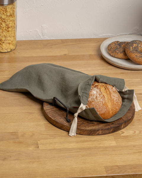 Loaf of bread in a stone green cloth bag on a wooden cutting board with a plate of bagels in the background.
