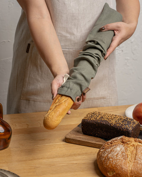Person holding a loaf of bread on a wooden table with apples and a bottle in the background