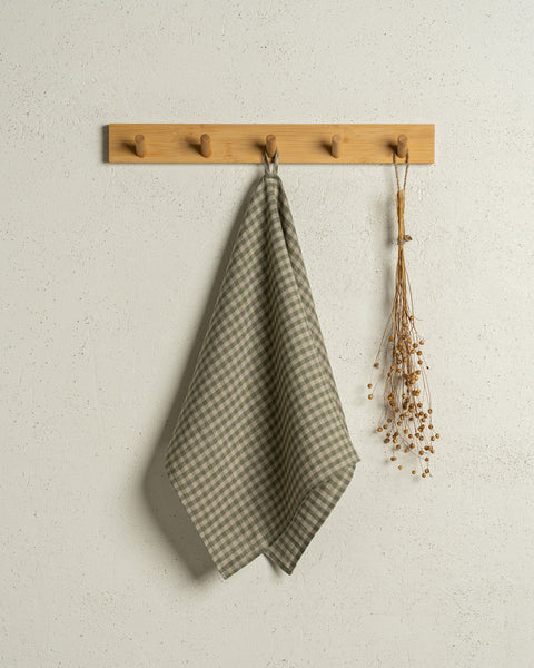 The menique linen kitchen dish towel in the color of green check hanging on a wooden rack on a wall.