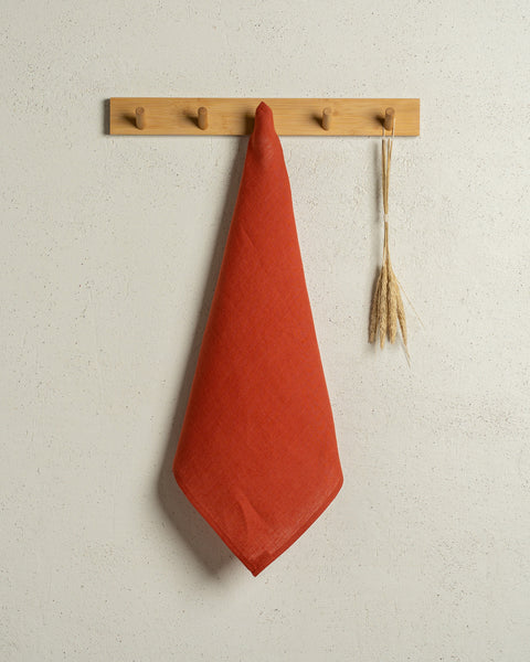 The menique square linen dish towel in color cinnamon red hanging on a wooden rack on a wall.