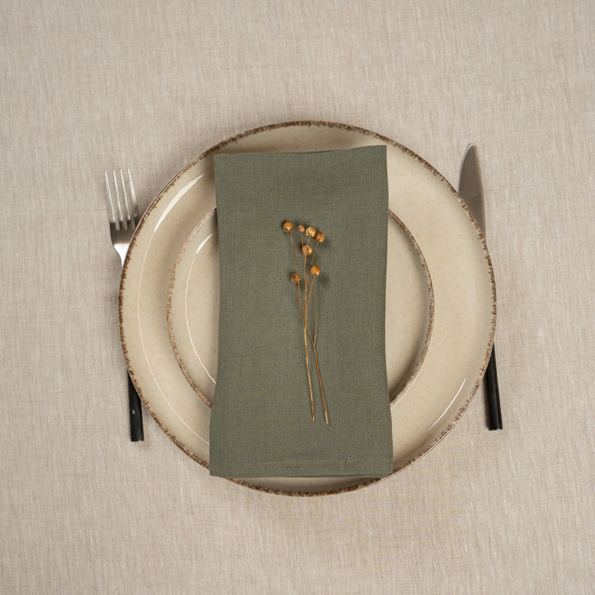 The menique linen napkin in spicy yellow color, put on top of two stacked plates . There are cutlery items by the sides of the plates. The background is a natural color linen tablecloth.