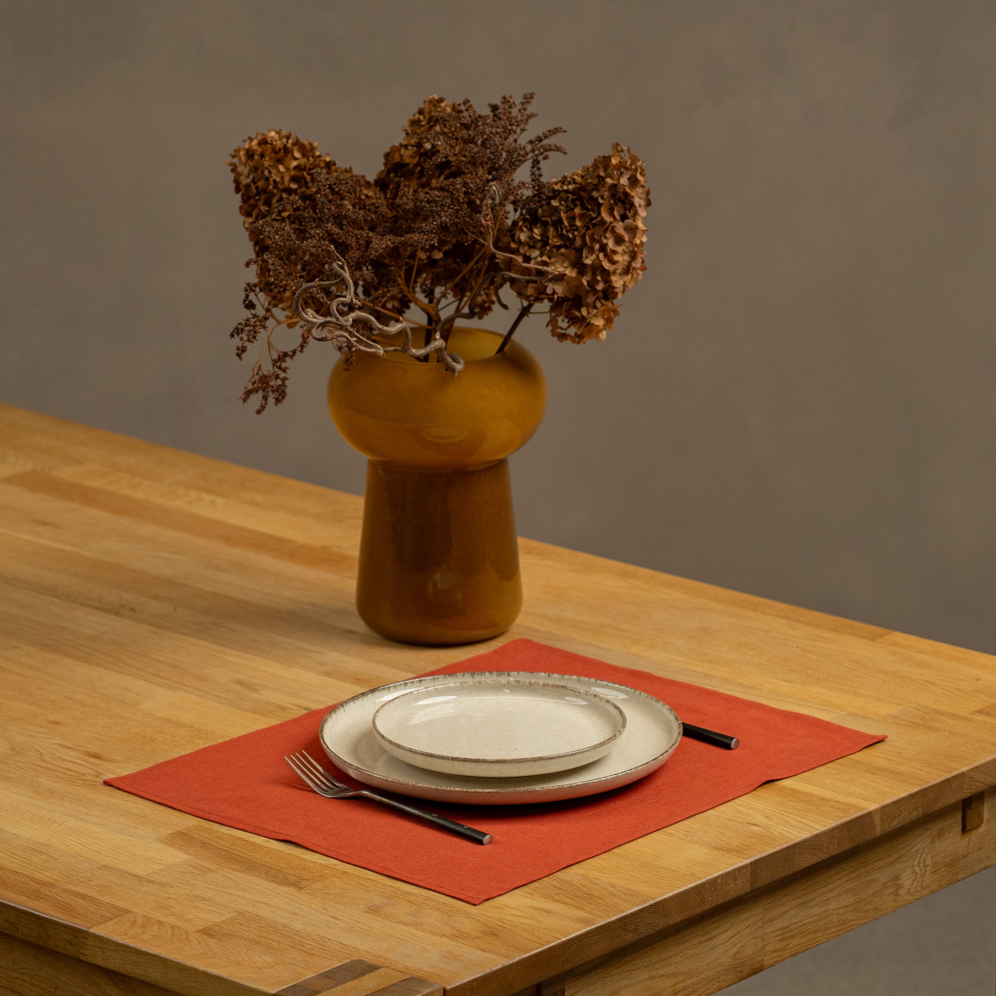 The menique linen placemat in natural color on a wooden table top. There is a vase on top of the table, as well as some cutlery by the sides of the plate, that is on top of the placemat.