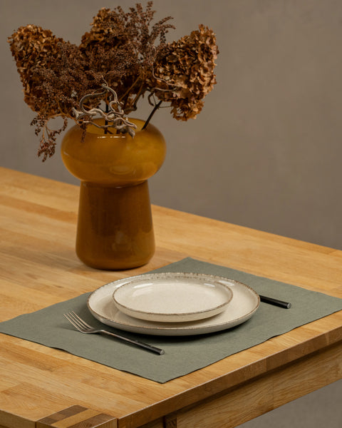The menique linen placemat in stone green color on a wooden table top. There is a vase on top of the table, as well as some cutlery by the sides of the plate, that is on top of the placemat.