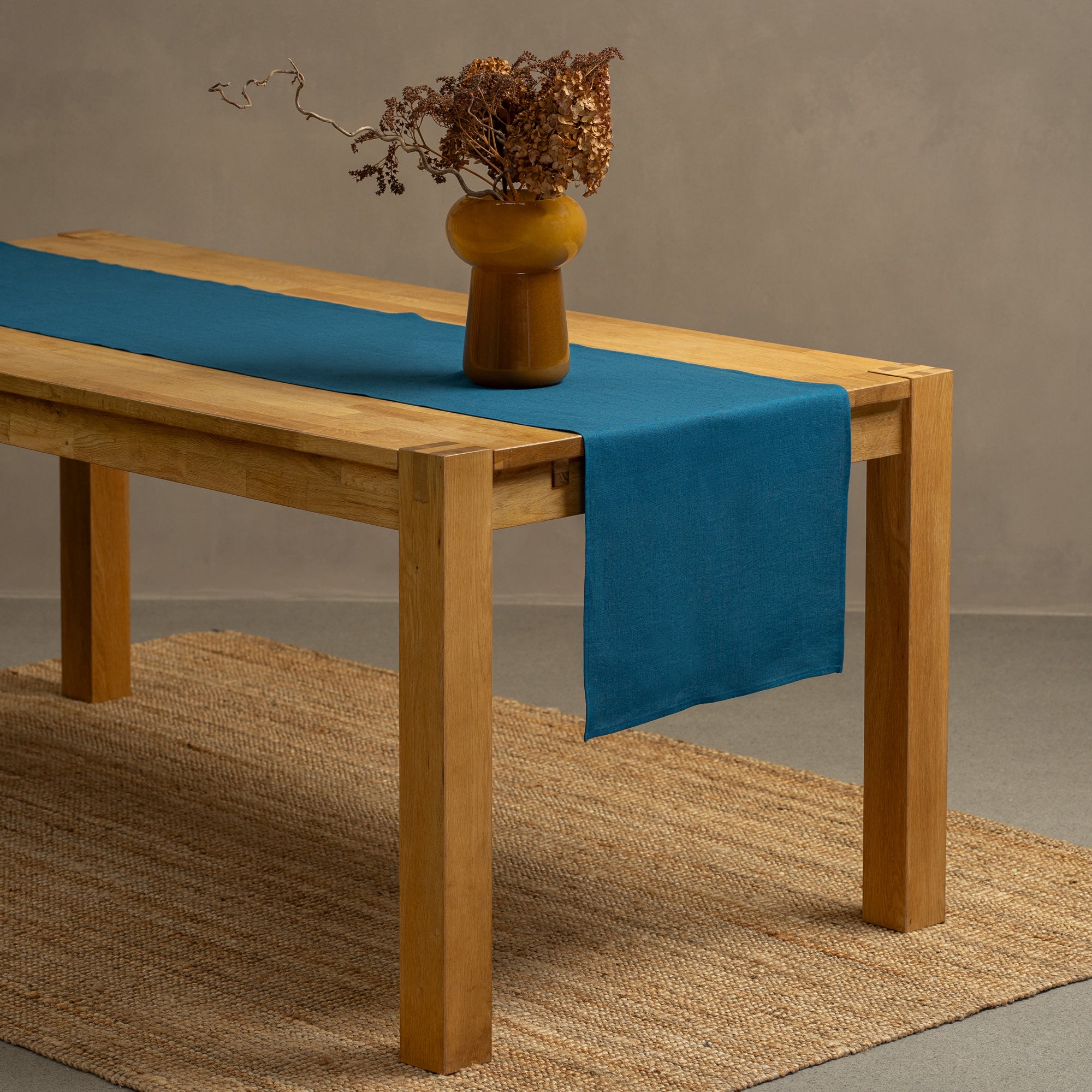 The menique linen table runner in dark green color, decorating a Christmas dinner table. There is a wine bottle, glasses, and a wooden table on top of the table top. there is also a white linen tablecloth underneath the runner.