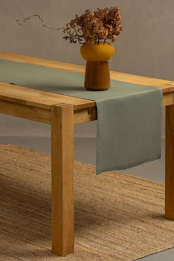 The stone green linen table runner on top of a wooden table, captured in a close up picture from the corner of the table. 