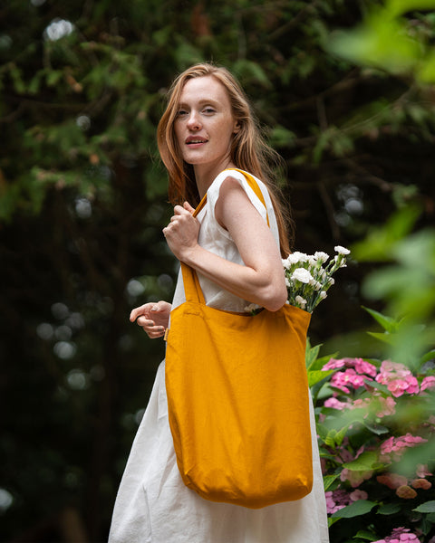 A fair-skinned woman with long, reddish-blonde hair looks over her shoulder with a gentle smile. She is wearing a light-colored dress and carrying a spicy yellow tote bag filled with white flowers. The background features blurred green foliage and pink flowers, suggesting an outdoor setting.