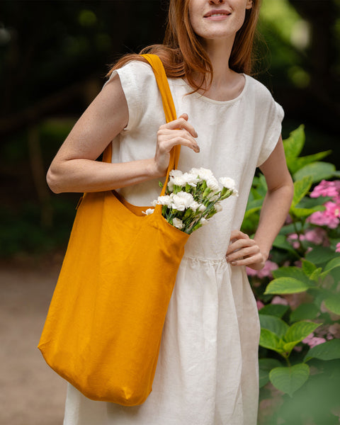A person with fair skin and long, reddish-brown hair smiles slightly while holding a spicy yellow linen tote bag filled with white flowers. They are wearing a light-colored dress and standing outdoors, with blurred green and pink foliage visible in the background.