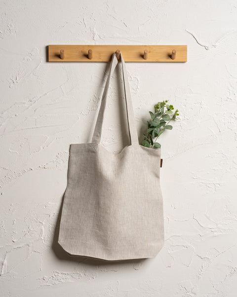 Menique reusable linen tote bag hangs from a wooden rack with four pegs mounted on a textured off-white wall. Green leafy stems are visible sticking out of the top of the bag.