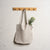 Menique natural color linen tote bag with long handles hangs from a branch of a light-colored tree. In the blurred background, there is a body of water reflecting the sky, surrounded by green grass and trees under a partly cloudy sky.