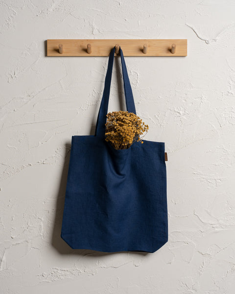 A storm blue reusable linen tote bag hangs from a wooden rack with four pegs mounted on a textured off-white wall. Dried yellow flowers are visible sticking out of the top of the bag.