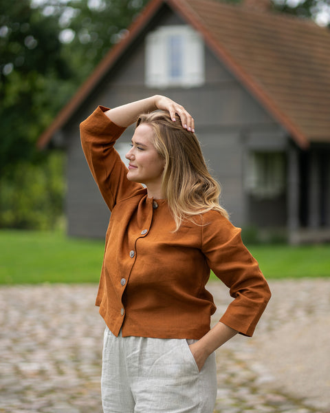 A woman with blonde hair poses outdoors wearing a rust-colored top and light trousers. She has one hand resting on her head with a traditional wooden house in the background.