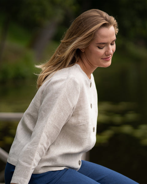 A young woman with long blonde hair sits outdoors wearing a light-colored sweater and dark blue jeans. She looks thoughtfully off to the side against a soft, blurred background of greenery and water.