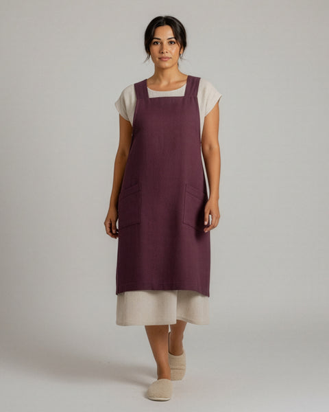 A woman wearing a shadow purple linen Japanese-style apron with front pockets, standing against a neutral backdrop over a cream dress and slippers.