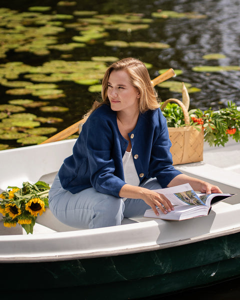 A blonde woman wearing a dark blue jacket over a light top sits in a rowboat leaning forward with a thoughtful expression. A basket of bright sunflowers is visible next to her against a backdrop of water and green trees.