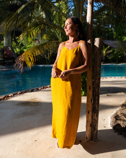 A woman with dark hair wearing a yellow sundress leans against a wooden post, looking to the left. Behind her is a bright blue pool surrounded by palm trees and tropical foliage. The ground is light-colored with shadows.