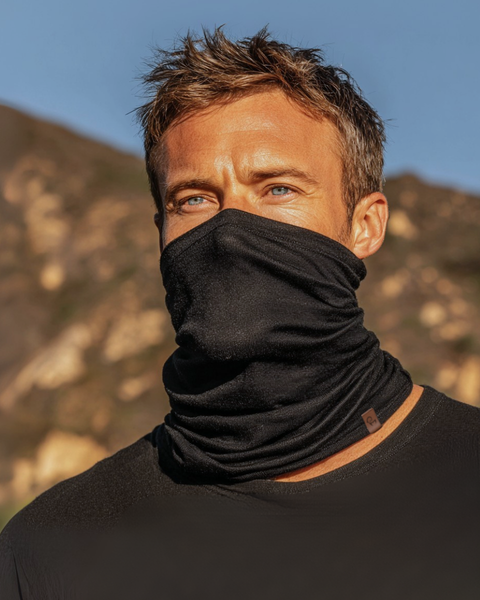 Man wearing a black Merino wool long neck gaiter covering his face, standing outdoors with mountains in the background.