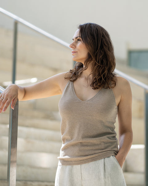 A woman with shoulder-length wavy brown hair stands in profile, looking to her left with one arm resting on a silver handrail and the other hand in her pocket. She is wearing a menique tank top and linen pants, and is positioned on light-colored stairs with a glass or clear barrier visible in the background.