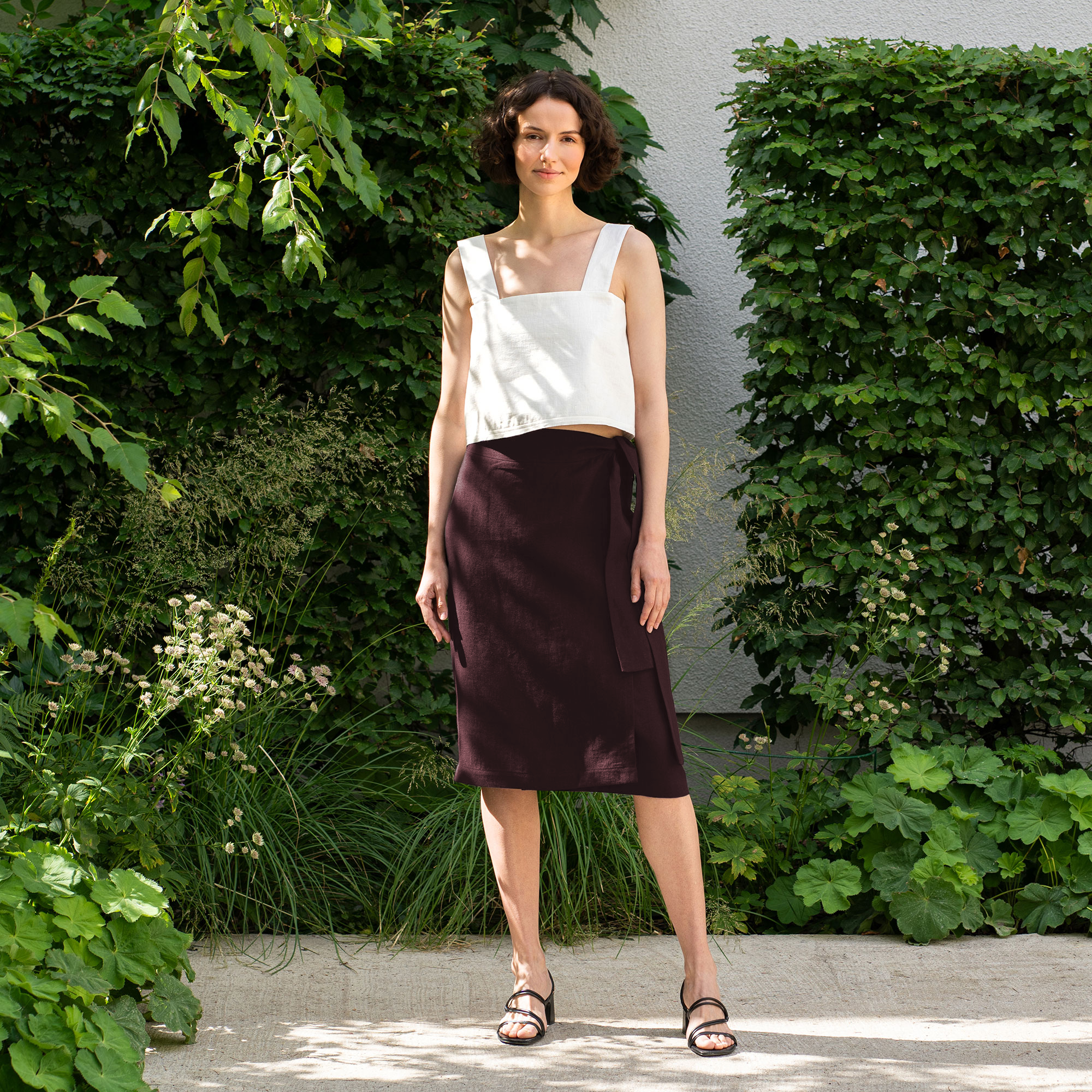 A woman with brown hair, wearing a natural linen button-front sleeveless top and a stone green wrap skirt, poses on outdoor concrete stairs.
