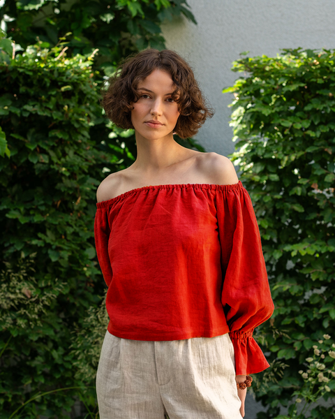 A woman with short, curly brown hair looks directly at the camera, standing in front of a green ivy-covered wall. She is wearing a pure red off-the-shoulder linen top with voluminous sleeves that gather at the wrists, and light-colored pants.