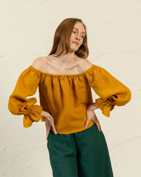 A light-skinned woman with long, straight auburn hair stands in front of a textured light-colored wall, looking slightly to her left. She is wearing a yellow off-the-shoulder top with voluminous sleeves gathered at the wrists and dark green pants. Her hands are on her hips.