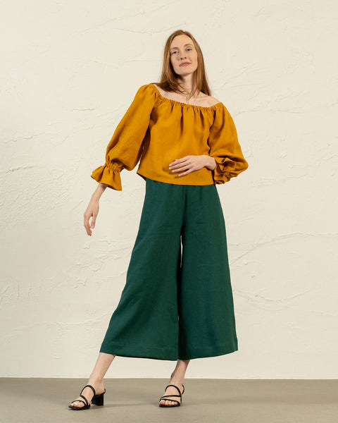 A light-skinned woman with long, straight auburn hair stands with one leg slightly bent, looking directly at the camera. She is wearing a yellow off-the-shoulder top with voluminous sleeves gathered at the wrists, dark green wide-leg culottes, and black open-toed block-heeled sandals. The background is a textured light-colored wall.